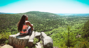 YOUNG WOMEN WITH LONG BLACK HAIR WEARING A COLORFUL PINK AND GRAY SLEEVELESS OUTFIT SITTING ON A A BIG STONE LOOKING OUT OVER GREEN HILLS