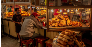 MAN IN A BIEGE LONGSLEEVED PULLOVER WEARING A RED HAT AT A SAN DIEGO PASTRY SHOP FOR TRAVELERS. YOUNG BOY EMPLOYEE OF BAKERY WEARING A MASK IS DRESSED IN BLACK 