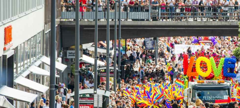 LARGE CROWD AT SWEDISH FESTIVAL WITH COLORFUL BANNER THAT SPELLS LOVE IN VIBRANT REDS, YELLOWS, GREENS, AND BLUES