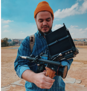 YOUNG MAN WEARING A RUST COLORED BEANIE HOLDING ELECTRONICS TRAVEL ESSENTIALS FOR HIS VACATION